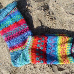 Könnte beinhalten: Eine farbenfrohe, handgestrickte Socke mit einem auffälligen Muster. Die Socke zeigt Bänder in Rot, Blau, Grün und Gelb mit aufwendigen geometrischen Designs. Der Bund und die Spitze sind einfarbig türkis. Die Socke liegt auf sandigem Untergrund.