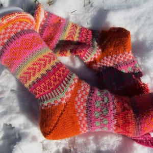 Könnte beinhalten: Ein Paar bunte Stricksocken mit einem lebendigen Muster aus Orange, Pink, Gelb und Grün. Die Socken liegen auf einer schneebedeckten Oberfläche und zeigen ihr aufwendiges Design und ihre warmen Farbtöne. Die Socken sind aus Wolle.