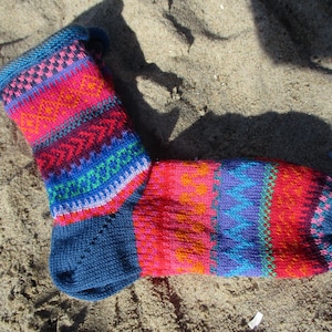 Könnte beinhalten: Eine farbenfrohe Stricksocke mit einem gemusterten Design. Die Socke hat einen blauen Bund, eine Ferse und eine Spitze mit Bändern in Pink, Orange, Rot, Blau und Grün. Die Socke liegt auf sandigem Untergrund.