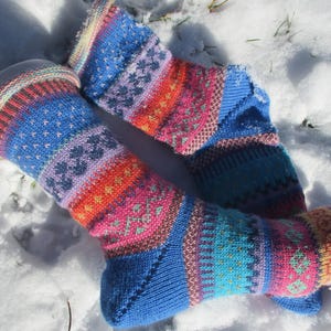 Könnte beinhalten: Ein Paar bunte Stricksocken mit einem lebendigen Muster. Die Socken haben eine blaue Basis mit Bändern in Orange, Rosa und Lila sowie aufwendige geometrische Designs. Die Socken liegen auf einer frischen weißen Schneedecke.