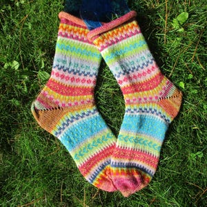 Könnte beinhalten: Ein Paar handgestrickte Socken mit einem lebendigen, mehrfarbigen Fair-Isle-Muster. Das Design umfasst horizontale Streifen in Pink, Blau, Gelb, Grün und Weiß mit komplizierten geometrischen Motiven im gesamten Gewebe und gerollten Bündchen.