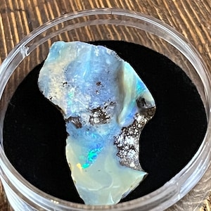 Könnte beinhalten: Ein roher Opal-Edelstein, der eine Mischung aus blauen, weißen und schillernden Farben zeigt. Der Opal befindet sich in einem transparenten Kunststoffbehälter mit schwarzem Innenraum, der die natürliche Schönheit des Steins hervorhebt. Der Stein hat eine raue, strukturierte Oberfläche.