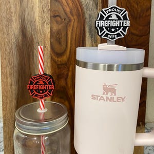 Peut inclure: Deux gobelets en acier inoxydable avec couvercles et pailles. Le gobelet de gauche est en verre transparent avec une paille rayée rouge et blanche. Le gobelet de droite est rose avec un logo blanc qui dit "STANLEY". Les deux gobelets ont un autocollant noir et blanc qui dit "PROUD FIREFIGHTER WIFE" avec un cœur rouge.