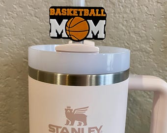 May include: A pink Stanley travel mug with a black and orange basketball mom straw topper. The mug has a white bear logo with a crown on its head and the word "STANLEY" printed below it.