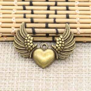 Könnte beinhalten: Ein goldfarbener Metallanhänger mit Herzform und zwei Flügeln. Die Flügel sind mit kleinen Punkten verziert und haben einen Vintage-Stil.