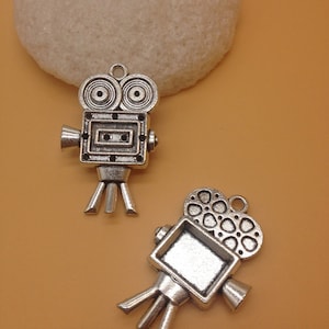 May include: Two silver-colored vintage movie camera charms. The charms have detailed designs, including lenses, a rectangular body, and tripod legs. The charms are on a light brown background, suggesting a theme related to cinema or filmmaking.