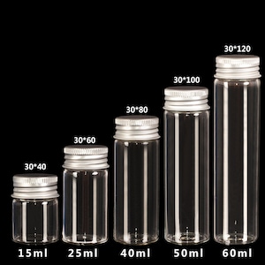 Op de afbeelding: Zes heldere glazen flessen met zilveren deksels in verschillende maten. De flessen zijn gelabeld met hun volume in milliliter: 15 ml, 25 ml, 40 ml, 50 ml en 60 ml. De flessen zijn gerangschikt op grootte, met de kleinste fles links en de grootste fles rechts.