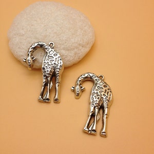 May include: Two silver-toned giraffe charms with a detailed design. The charms are hanging from a white surface.
