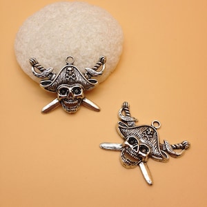 May include: Two silver-toned pirate skull charms with crossed swords and a pirate hat. The charms are on a light brown background.