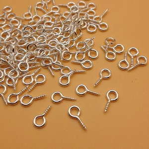 Può includere: Un mucchio di ganci a occhiello a vite di colore argento. I ganci hanno un piccolo anello nella parte superiore e una base a vite.