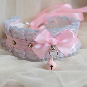 Może przedstawiać: Delikatny, jasnoniebieski choker z koronki z różową wstążką satynową i małym, różowym dzwonkiem.