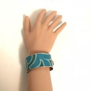 May include: A wide, turquoise enamel bracelet with a silver-tone metal base. The bracelet features a leaf-like design in shades of turquoise and teal. The bracelet is approximately 3.8 cm wide.