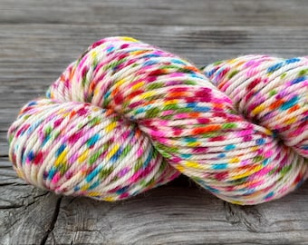 May include: A skein of twisted yarn featuring a cream-colored base with a vibrant, multi-colored speckled pattern. The yarn displays small, distinct flecks of bright pink, yellow, blue, and green distributed evenly throughout the twisted fibers.