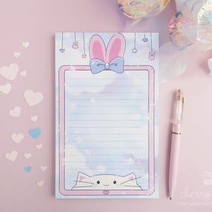May include: A lined notepad with a bunny ear and bow design, a cat graphic, and a pink border. A pink pen rests beside the notepad on a pink surface. Decorative hearts are scattered around the notepad.