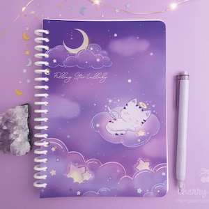 May include: A spiral-bound notebook with a purple cover featuring a sleeping cat angel on a cloud, surrounded by stars and a crescent moon. The text "Falling Star Lullaby" is written on the cover. A white pen rests beside the notebook.