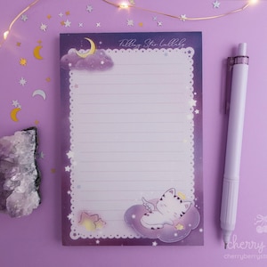 May include: A lined notepad with a celestial theme, featuring a sleeping cat on a cloud, a crescent moon, and stars. A white pen rests beside the notepad. The notepad has the text "Falling Star Lullaby".