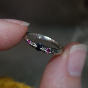 May include: A silver ring with a textured band and three gemstones. The ring has one black gemstone and two pink gemstones.