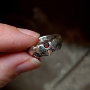 Könnte beinhalten: Ein silberner Ring mit einem strukturierten, organischen Design. Der Ring hat einen kleinen, runden, roten Edelstein, der in das Band eingesetzt ist. Der Ring wird von den Fingern einer Person gehalten.