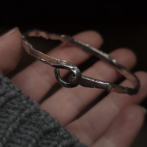 May include: A silver-toned bangle bracelet with a hammered texture and a hook-and-eye closure. The bracelet is held in a hand, showcasing its simple, yet elegant design. The metal has a slightly tarnished appearance, adding to its rustic charm.