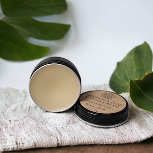 May include: Open black tin of solid perfume with a light beige color, next to the closed tin. The lid of the closed tin has a label that reads "Magnolia Leaf & Tonka Bean Solid Perfume". The perfume is on a white cloth.