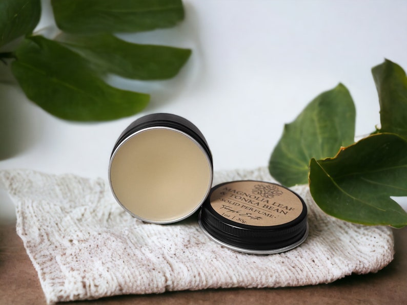 May include: Open black tin of solid perfume with a light beige color, next to the closed tin. The lid of the closed tin has a label that reads "Magnolia Leaf & Tonka Bean Solid Perfume". The perfume is on a white cloth.