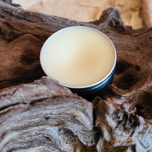 Pu&ograve; includere: Un piccolo contenitore rotondo di latta con un coperchio, riempito di un balsamo giallo chiaro. Il contenitore &egrave; appoggiato su un pezzo di legno di deriva consumato.