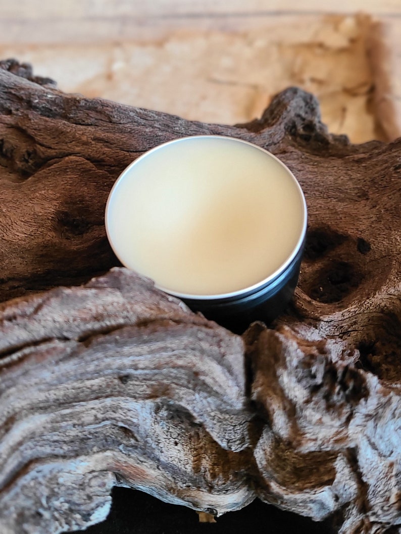 Pu&ograve; includere: Un piccolo contenitore rotondo di latta con un coperchio, riempito di un balsamo giallo chiaro. Il contenitore &egrave; appoggiato su un pezzo di legno di deriva consumato.