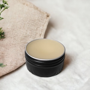 May include: A small, round black tin with a silver rim, filled with a light beige balm. The tin sits on a white surface next to a beige linen cloth and green sprigs. The balm appears smooth and creamy.