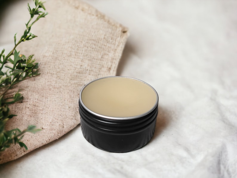 May include: A small, round black tin with a silver rim, filled with a light beige balm. The tin sits on a white surface next to a beige linen cloth and green sprigs. The balm appears smooth and creamy.