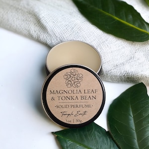 May include: An open, round, metal tin of solid perfume with the lid beside it. The lid is brown with the text "MAGNOLIA LEAF & TONKA BEAN SOLID PERFUME" and a floral design. The tin contains 1 oz. of perfume.
