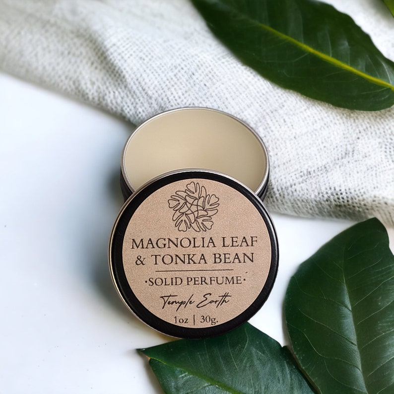 May include: An open, round, metal tin of solid perfume with the lid beside it. The lid is brown with the text "MAGNOLIA LEAF & TONKA BEAN SOLID PERFUME" and a floral design. The tin contains 1 oz. of perfume.
