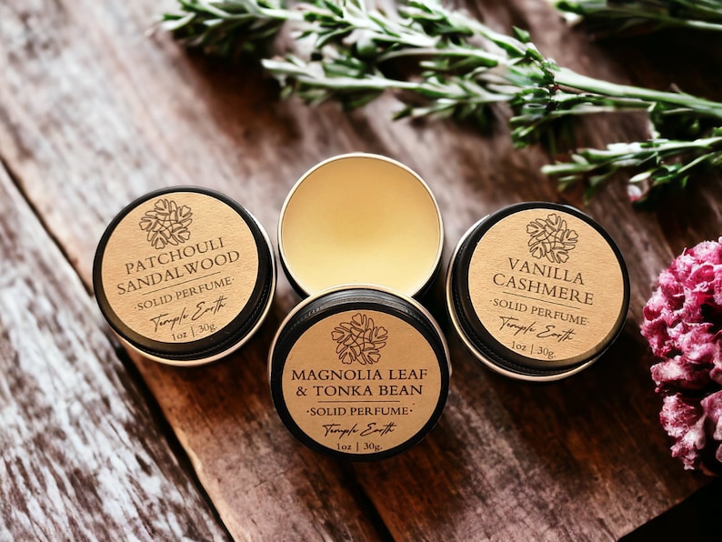 May include: Four small, round solid perfume tins with black rims and gold lids are arranged on a wooden surface. The labels read "Patchouli Sandalwood", "Vanilla Cashmere", and "Magnolia Leaf & Tonka Bean". The open tin reveals the solid perfume.