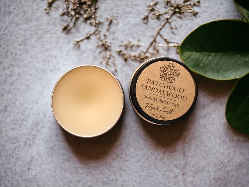 May include: Two small, round containers of solid perfume. One is open, revealing a light beige balm. The other is closed, with a brown label that reads "Patchouli Sandalwood Solid Perfume" and "Temple Earth 1oz | 30g".