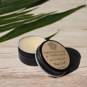 May include: Open black tin of solid perfume with a light beige balm. The lid, slightly ajar, displays the text "Magnolia Leaf & Tonka Bean Solid Perfume" and "Temple Earth". A green leaf is in the background. The perfume is 1 oz / 30g.