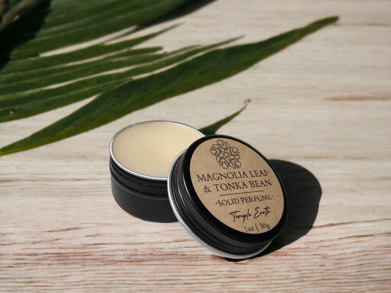 May include: Open black tin of solid perfume with a light beige balm. The lid, slightly ajar, displays the text "Magnolia Leaf & Tonka Bean Solid Perfume" and "Temple Earth". A green leaf is in the background. The perfume is 1 oz / 30g.
