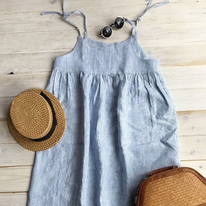 May include: A blue and white striped linen dress with thin straps. The dress has a loose fit and a gathered waist. It is photographed on a white wooden background with a straw hat and a woven bag.