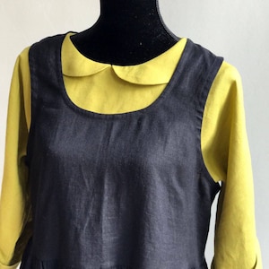 May include: A black linen dress with a gathered skirt and a Peter Pan collar. The dress is layered over a yellow collared shirt.