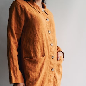 May include: A long, loose-fitting, mustard yellow linen shirt with a collar and button closure. The shirt has a single patch pocket on the left side and a slit in the back.