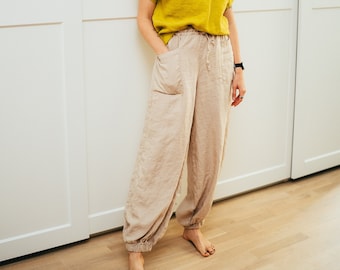 May include: A woman wearing a yellow linen top and beige linen pants with pockets. She is standing in front of a white wall.