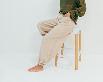 May include: A woman wearing a green, textured, long-sleeved shirt and beige, wide-leg pants sits on a white stool with wooden legs. She is smiling and looking to the right.