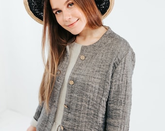 May include: A woman wearing a gray linen jacket with a black and brown straw hat. The jacket has a button-up front and long sleeves.