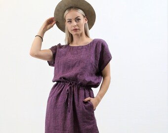 May include: A purple linen dress with a drawstring waist and short sleeves. The dress is knee-length and has a relaxed fit. The model is wearing a brown hat and white sneakers.