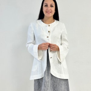 May include: A white linen jacket with a round neckline and long sleeves. The jacket has a button closure and a slightly flared hem. The model is wearing the jacket over a gray linen dress.