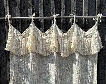 May include: A white cotton curtain with a ruffled top and a textured, crinkled fabric. The curtain is hanging from a silver rod against a dark wood background.