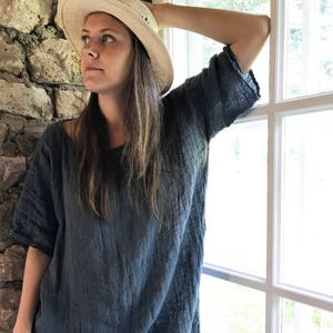 Op de afbeelding: Een vrouw draagt een loszittende, donkerblauwe linnen tuniek met een brede, ronde halslijn. Ze draagt een strohoed en staat bij een raam.