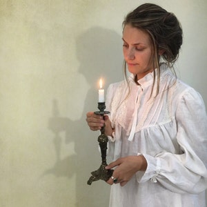 Pu&ograve; includere: Una donna in abito bianco tiene in mano una candela accesa in un candelabro antico in ottone. La fiamma della candela brilla intensamente, proiettando una luce calda. Il candelabro ha un design ornato.