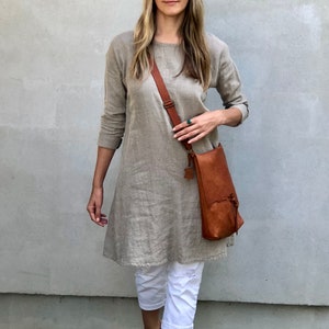 May include: A woman wearing a light brown linen dress with a brown leather crossbody bag. She is wearing white pants and brown sandals. The woman is standing in front of a gray wall.