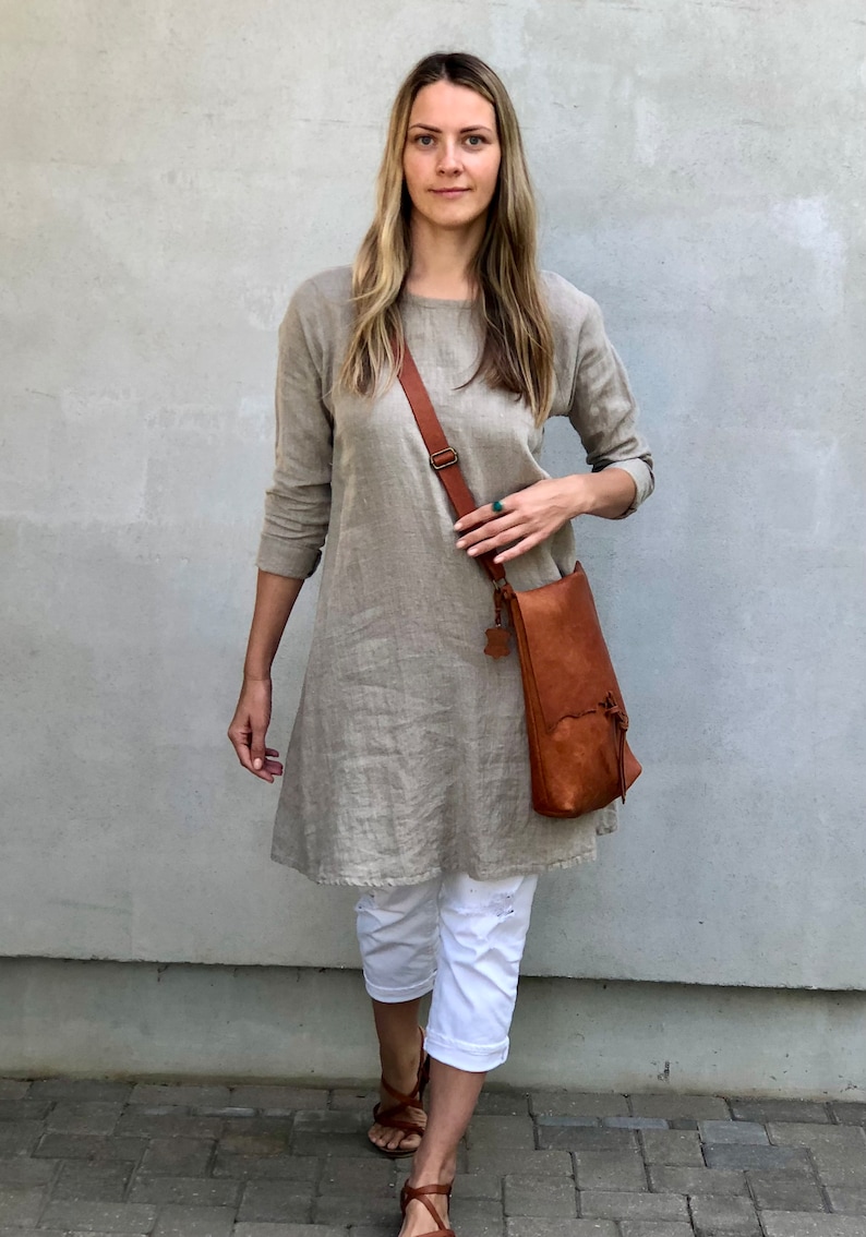 May include: A woman wearing a light brown linen dress with a brown leather crossbody bag. She is wearing white pants and brown sandals. The woman is standing in front of a gray wall.