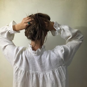 Pu&ograve; includere: Una camicetta di lino bianca con maniche lunghe e polsini a balze. La camicetta ha un colletto alto a balze e un corpetto arricciato. La persona indossa un anello con una pietra turchese. L'immagine &egrave; ripresa da dietro.