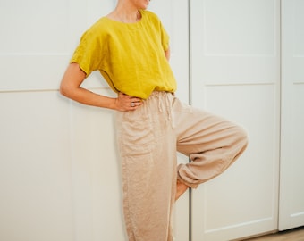 May include: A woman wearing a mustard yellow linen top and beige linen pants. The top has short sleeves and a round neckline. The pants have a drawstring waist and pockets. The woman is standing on one leg, leaning against a white door.